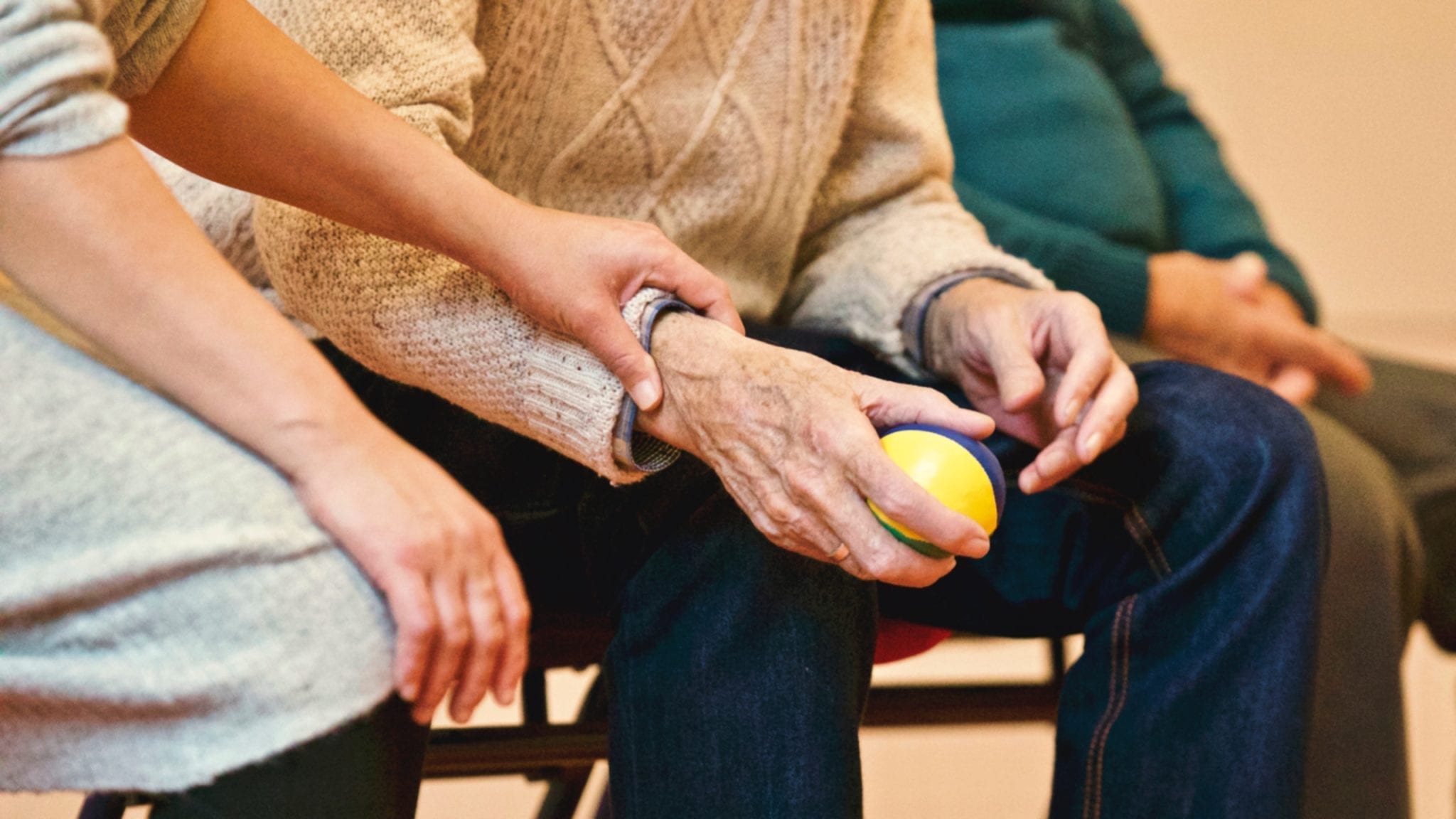 An elderly man sits holding a ball while another person gently grabs his wrist