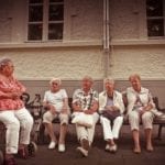 A group of female nursing home residents sit outside on a bench. One woman sits on a walker.