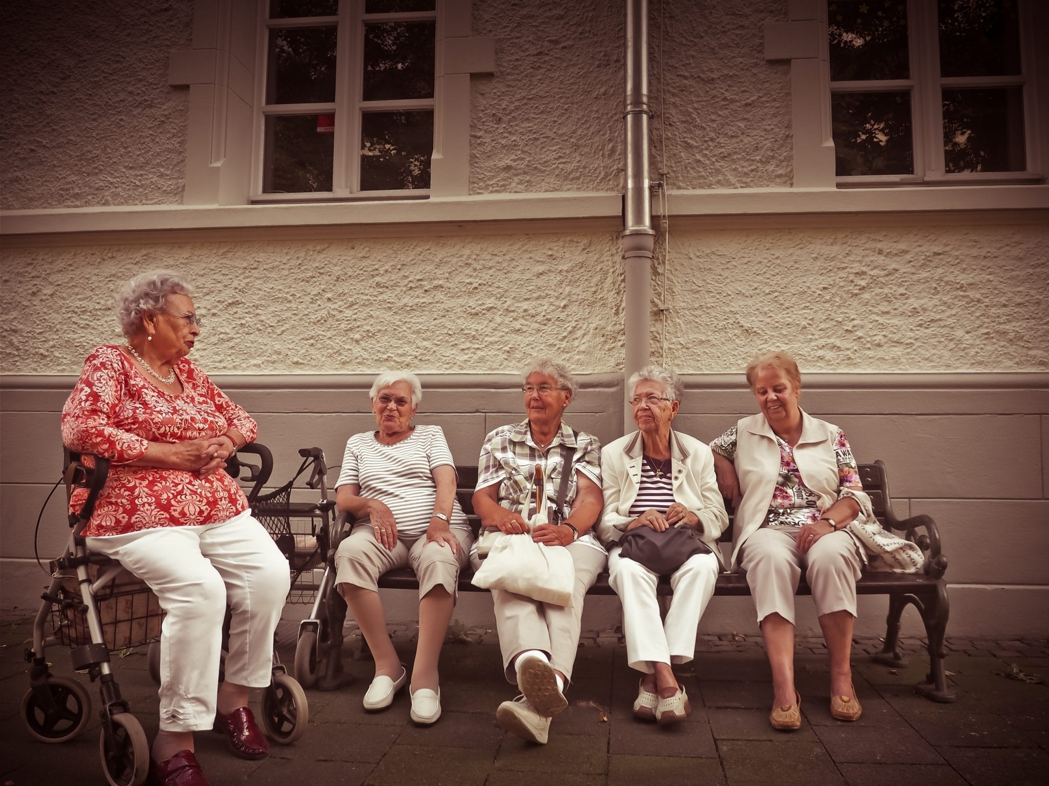 A group of female nursing home residents sit outside on a bench. One woman sits on a walker.