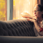 an older woman with glasses sadly sits on a couch