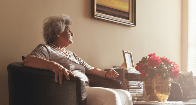 An older woman sits and stares out a window with a look of concern.