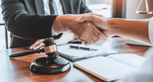 A lawyer shakes hands with a client while sitting at a desk. A brown gavel sits on the desk in the foreground.