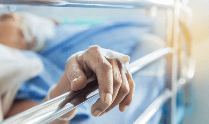 An older person in a hospital bed rests their hand on a guard rail