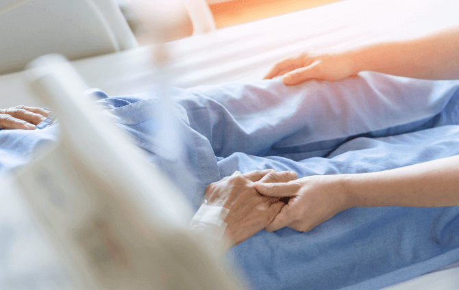 an older person sits in a hospital bed holding a younger person's hand