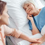 a female caregiver sits next to an older woman who is lying in bed