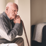 An older man sits sadly and rests his hands on a cane