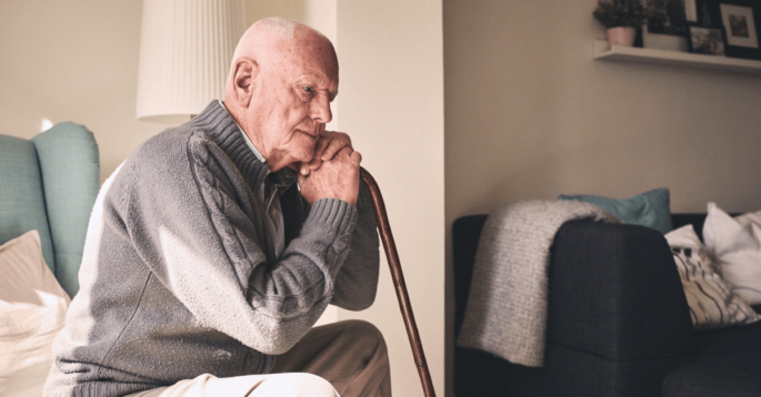 An older man sits sadly and rests his hands on a cane
