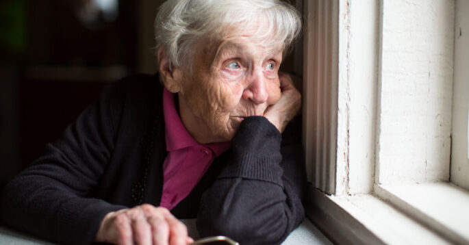 an elderly woman stares out a window and looks concerned