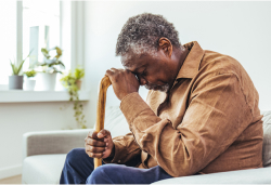 An older man sitting on a couch with his head resting on a cane, appearing distressed.
