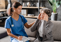 A medical professional comforting an older woman who is holding her head in pain during a nursing home visit.