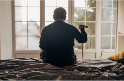 An older man sitting on a bed with a cane, looking out a window in a nursing home.