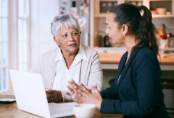 A nursing home lawyer with a laptop speaks with a client at a kitchen table.