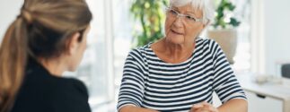 An older woman with white hair and glasses sits at a table speaking seriously with a younger woman, possibly discussing a sensitive issue such as nursing home abuse