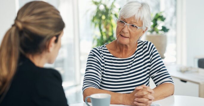 An older woman with white hair and glasses sits at a table speaking seriously with a younger woman, possibly discussing a sensitive issue such as nursing home abuse
