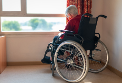 An elderly woman in a wheelchair sits by a nursing home window looking outside.