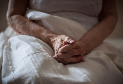 An elderly woman sits in bed with her hands folded on a blanket.