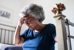 Older woman holding her head in discomfort while sitting on the edge of a bed.