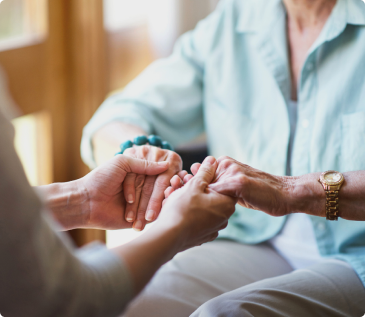A caregiver gently holds the hands of an older adult during a seated conversation.