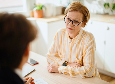 An older adult sits at a table with arms folded, looking concerned during a conversation with someone seated across from them.