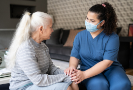 A caregiver wearing a mask sits with an older adult, holding their hands during a conversation.