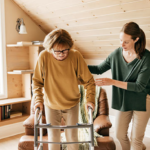 A caregiver walks beside an older adult using a walker in an assisted living community.