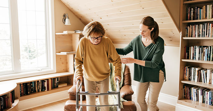 A caregiver walks beside an older adult using a walker in an assisted living community.