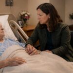 A woman holds the hand of an older adult lying in a hospital bed.