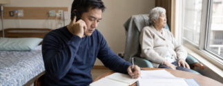 A man talks on the phone and takes notes while an older adult sits by a window in a nursing home room.