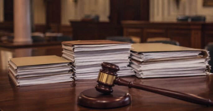 Stacks of legal documents sit on a courtroom table beside a judge's gavel.