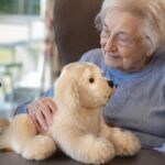 An older adult cares for a robot pet for dementia patients while a caregiver sits beside her.