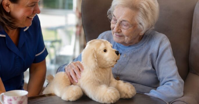 An older adult cares for a robot pet for dementia patients while a caregiver sits beside her.