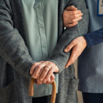 A caregiver supports an older adult using a cane in a nursing home.