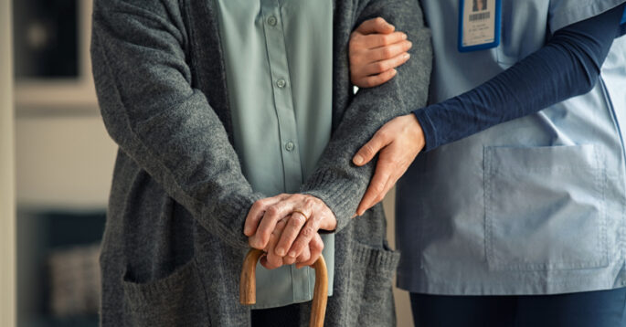 A caregiver supports an older adult using a cane in a nursing home.
